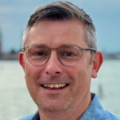 Close-up headshot of a person wearing rectangular eyeglasses and a light blue collared shirt, facing the camera. The background is softly blurred, showing a body of water and an overcast sky. The image is well lit with the person centered in the frame.