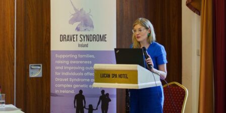 A women in a blue jumpsuit, standing behind a podium to deliver a talk.