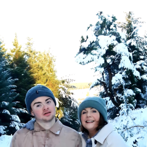 A mother and son smiling at the camera, standing in front of snow covered trees.