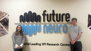 A woman and a man standing on either side of a large blue and navy FutureNeuro sign on the wall.