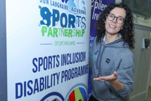 A woman wearing glasses and a grey hoodie standing in front of the DLR Sports Partnership pull-up banner.