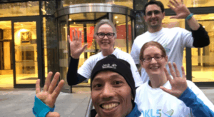 Four people, three females and 1 male, wearing white t-shirts and waving.