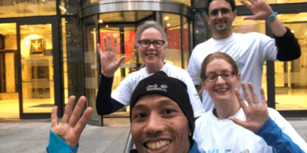 Four people, three females and 1 male, wearing white t-shirts and waving.