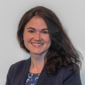 A headshot of a woman with thick dark hair, who is smiling and wearing a navy blazer.
