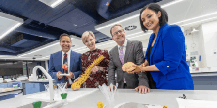 Four people stand smiling in a bright, modern laboratory. They each hold anatomical models — a heart, a spine, and a brain. Lab equipment and benches are in the foreground, and overhead lighting and vents are visible above them.