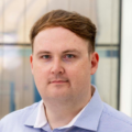 Professional headshot of a person wearing a light blue patterned collared shirt, facing the camera. The image is cropped from the shoulders up, with the subject centered in the frame. The background is softly blurred and appears to be an office environment with glass panels and neutral tones.