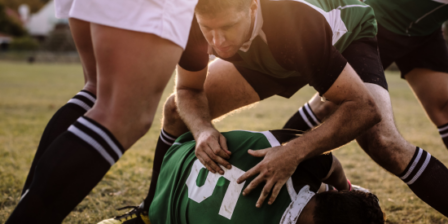 A rugby player in a green jersey is tackled to the ground during a match, with an opposing player in white leaning over him. Other players' legs are visible in the background on a grass field.