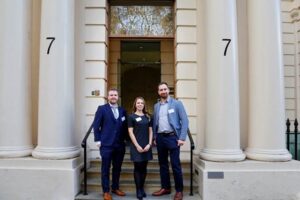 Three professionally dressed researchers standing together on the steps outside a classical building with tall columns and the number 7 displayed on either side of the entrance.