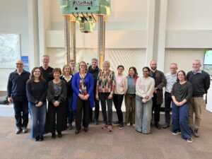 A group of fifteen people standing together in two rows inside a large indoor space. Behind them is a tall industrial machine with metal columns. The group is centered in the frame, standing on a brown floor with light‑coloured walls and display panels in the background. Everyone is facing the camera and standing closely together.