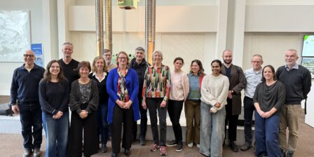 A group of fifteen people standing together in two rows inside a large indoor space. Behind them is a tall industrial machine with metal columns. The group is centered in the frame, standing on a brown floor with light‑coloured walls and display panels in the background. Everyone is facing the camera and standing closely together.