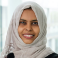 Head-and-shoulders portrait of a person wearing a light-coloured headscarf, looking directly at the camera, with a softly blurred indoor background.