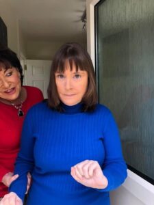Two women standing in a doorway or hallway. The woman in the foreground has shoulder-length brown hair with bangs and is wearing a bright blue ribbed turtleneck sweater, with her hands clasped in front of her. Behind her, partially visible, is an older woman wearing a red top and a dark pendant necklace. 