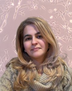 A person with long, light‑coloured hair sits facing the camera, wearing a thick, cable‑knit sweater. The background shows a textured pattern resembling abstract, pale shapes on a soft pink surface.