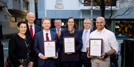Group of seven people standing outside the RCSI building, with three people in the front row holding framed Vice Chancellor Innovation Awards 2025 certificates. The group is posed on a city street in front of a modern glass and stone building displaying the RCSI logo.