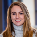 A headshot of a person with long light-brown hair and blue eyes, wearing a light grey turtleneck sweater. The person is centered in the frame, facing the camera with a slight smile. The background is softly blurred, showing indoor tones of blue and neutral colours.