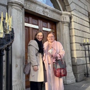 Two women standing outside the entrance of a historic stone building with large wooden doors framed by decorative columns. One woman wears a light beige coat and crossbody bag, while the other wears a pale pink coat and carries a red-and-grey handbag. Black iron railings with gold-tipped finials line the foreground, and sunlight casts shadows across the stone façade. Small plaques are visible above the doorway, indicating offices within the building.