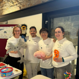Four people wearing white FamilyGenomics Research Group t-shirts stand together at an outreach event display. The group is holding educational models and toys, including a plush grey animal, a model brain, an orange anatomical model, and a black-and-yellow bird puppet. A research poster about the FamilySleeps study is displayed on a noticeboard in the background, while tables in the foreground contain activity materials and demonstration items. The event is taking place indoors against a stone wall and glass door backdrop