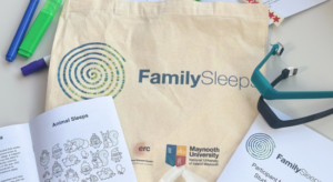 Cream-colored canvas tote bag printed with the FamilySleeps logo and branding, photographed on a white surface alongside study materials. Around the bag are colorful markers, a booklet titled “Animal Sleeps” with illustrated animals, a participant information leaflet, and a teal wearable sleep-monitoring device with a charging cable. The Maynooth University and ERC logos are visible on the tote bag, indicating participation in a sleep research study.