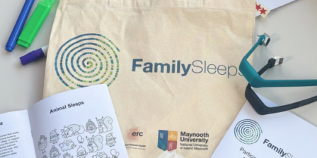 Cream-colored canvas tote bag printed with the FamilySleeps logo and branding, photographed on a white surface alongside study materials. Around the bag are colorful markers, a booklet titled “Animal Sleeps” with illustrated animals, a participant information leaflet, and a teal wearable sleep-monitoring device with a charging cable. The Maynooth University and ERC logos are visible on the tote bag, indicating participation in a sleep research study.