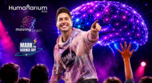 Promotional graphic featuring a presenter in a pink and purple hoodie pointing toward the audience on a brightly lit stage. Behind the presenter is a large glowing digital brain illustration in purple, blue, and pink tones, with audience silhouettes visible in the foreground, including a raised hand. Logos for Humanarium RCSI, My Moving Brain, and Mark the Science Guy appear on the left side against a vibrant purple background.