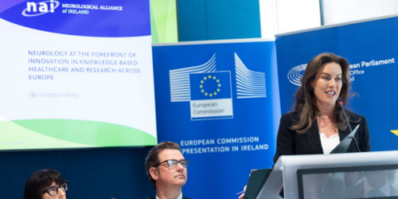 The Minister of Health speaks at a podium during a conference event, with European Commission and Neurological Alliance of Ireland banners displayed behind the stage. Two seated panel members are visible in the foreground, while the Minister addresses the audience from a lectern equipped with a microphone. The setting is a formal healthcare and research-focused event with European institutional branding.