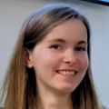 Close-up headshot of a person with long, straight hair, facing the camera and showing teeth in a smile. The person is indoors against a light-colored, slightly blurred background, with even lighting across the face. Only the head and upper shoulders are visible.