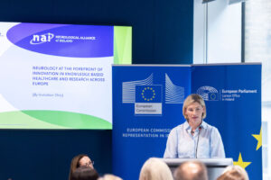Nina Carberry MEP speaks at a podium during a Neurological Alliance of Ireland event focused on innovation, healthcare, and neurological research in Europe. European Commission and European Parliament Liaison Office in Ireland banners are displayed behind the speaker, alongside a presentation screen showing the event title. Audience members are visible in the foreground in a formal conference setting.