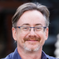 Close-up head-and-shoulders portrait of a person wearing blue-framed glasses and a blue collared shirt. The person has short gray hair and facial hair. The background is blurred and dark, placing focus on the face.