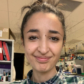 Close-up portrait of a person looking at the camera in an indoor laboratory or research workspace. The person is wearing a green and blue plaid shirt, with hair tied up. Shelves with red binders, laboratory equipment, and various containers are visible in the background.