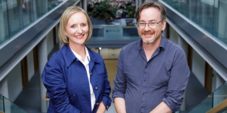 Two people stand side by side on an indoor staircase in a modern office atrium with glass railings and upper-level walkways. One person wears a blue jacket over a white shirt, and the other wears a patterned blue button-down shirt and jeans. The bright, contemporary workspace features natural light and greenery in the background.