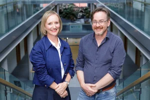 Two people stand side by side on an indoor staircase in a modern office atrium with glass railings and upper-level walkways. One person wears a blue jacket over a white shirt, and the other wears a patterned blue button-down shirt and jeans. The bright, contemporary workspace features natural light and greenery in the background.