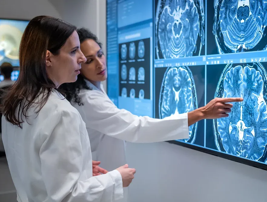 Two healthcare professionals in white coats look at an MRI scan of a patient's brain. One of the women is pointing out something on the scan to the other.