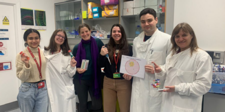 Six colleagues pose together in a laboratory, some wearing lab coats and holding research materials, including sample tubes, a small model, and a hand-drawn scientific illustration.